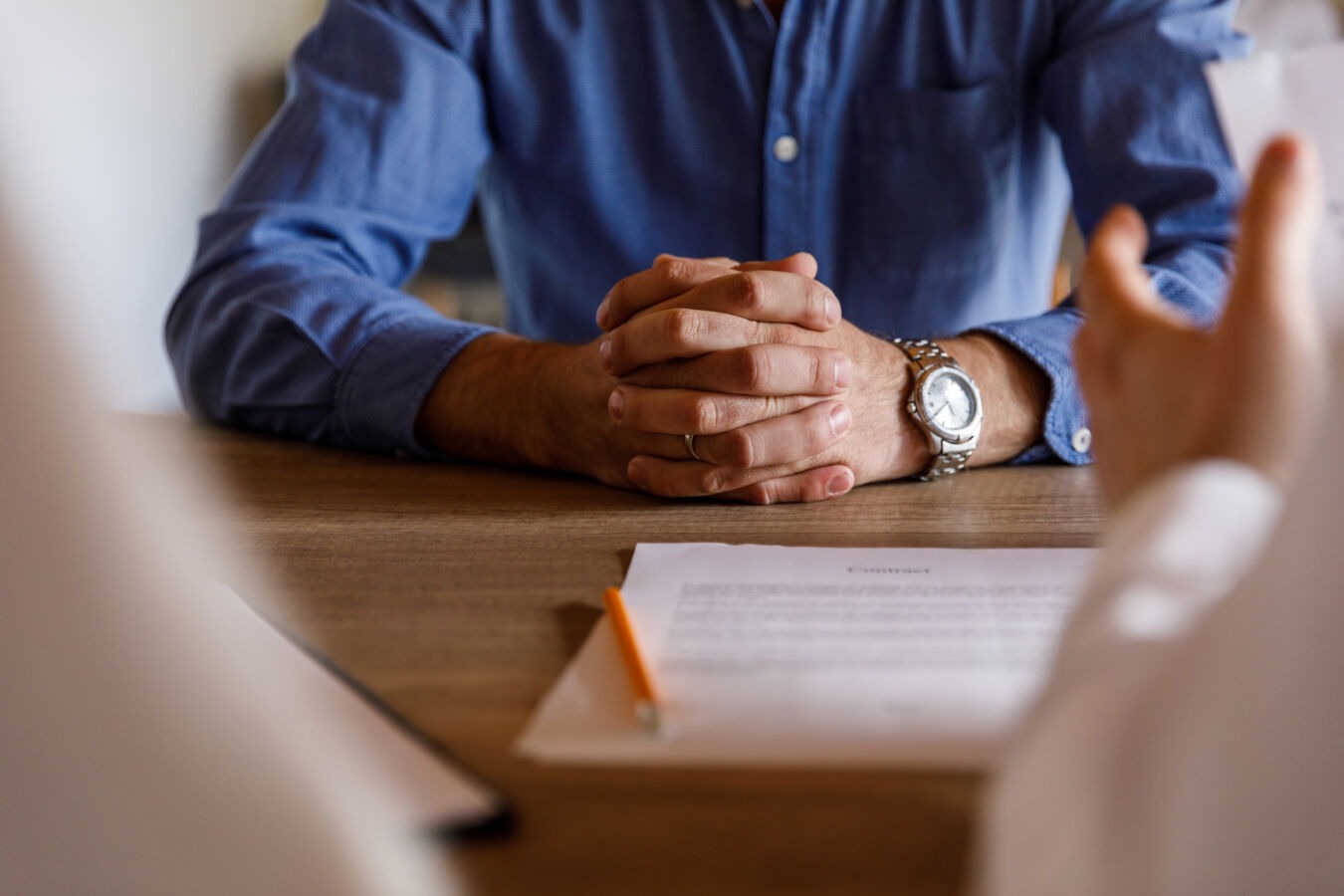 Man with clasped hands sitting across the table from someone speaking
